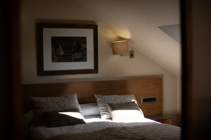 Made bed with a sunlit corner, framed art and a wall sconce above
