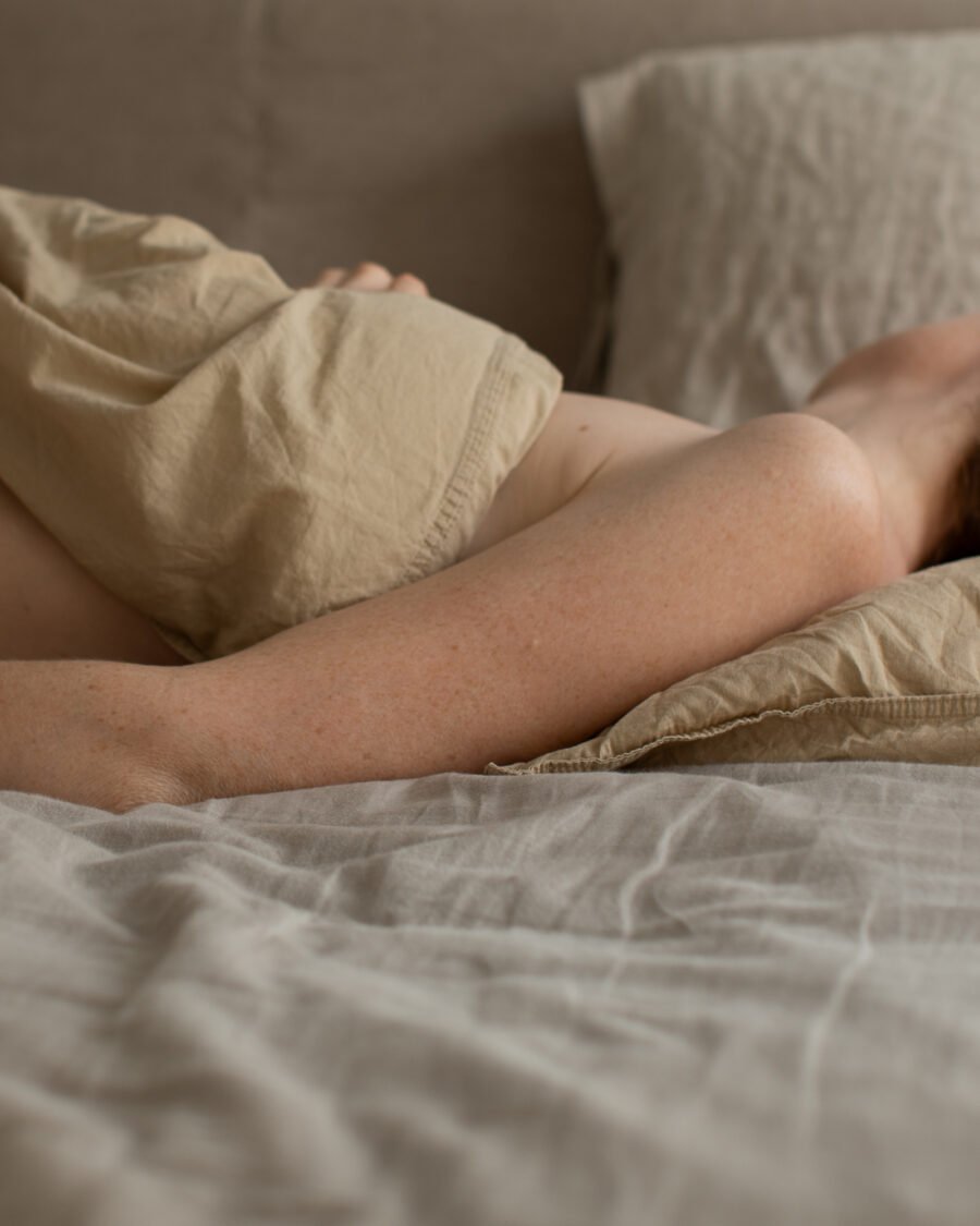 Woman sleeping face-down in bed, pulling a linen sheet over her head in soft morning light